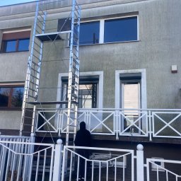 Jarek Wachowiak - Pracownik w czarnej bluzie stoi na balkonie zabezpieczonym białą balustradą, przed budynkiem z szarą elewacją, na tle rozstawiona aluminiowa drabina rusztowaniowa.