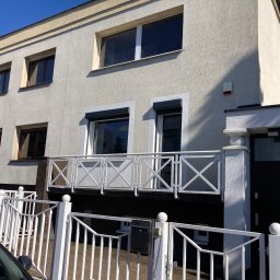 Jarek Wachowiak - Dwupiętrowy budynek z jasną elewacją, balkonem z białą balustradą i białym ogrodzeniem, widziany z poziomu ulicy, z fragmentem samochodu w kadrze.