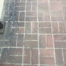 Jarek Wachowiak - Ceglana kostka brukowa, część wyczyszczona z ciemnych zabrudzeń, widoczny fragment buta i kratki ściekowej, ujęcie z góry.