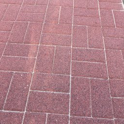 Jarek Wachowiak - Czerwona kostka brukowa ułożona w regularny wzór. Widoczne fugi wypełnione piaskiem. Fragment trawnika w prawym górnym rogu.