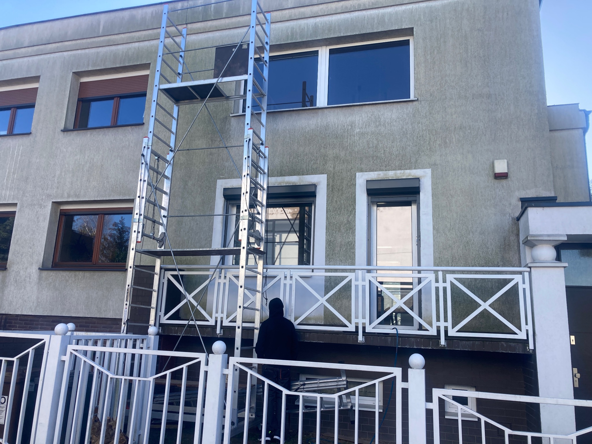 Pracownik w czarnej bluzie stoi na balkonie zabezpieczonym białą balustradą, przed budynkiem z szarą elewacją, na tle rozstawiona aluminiowa drabina rusztowaniowa.