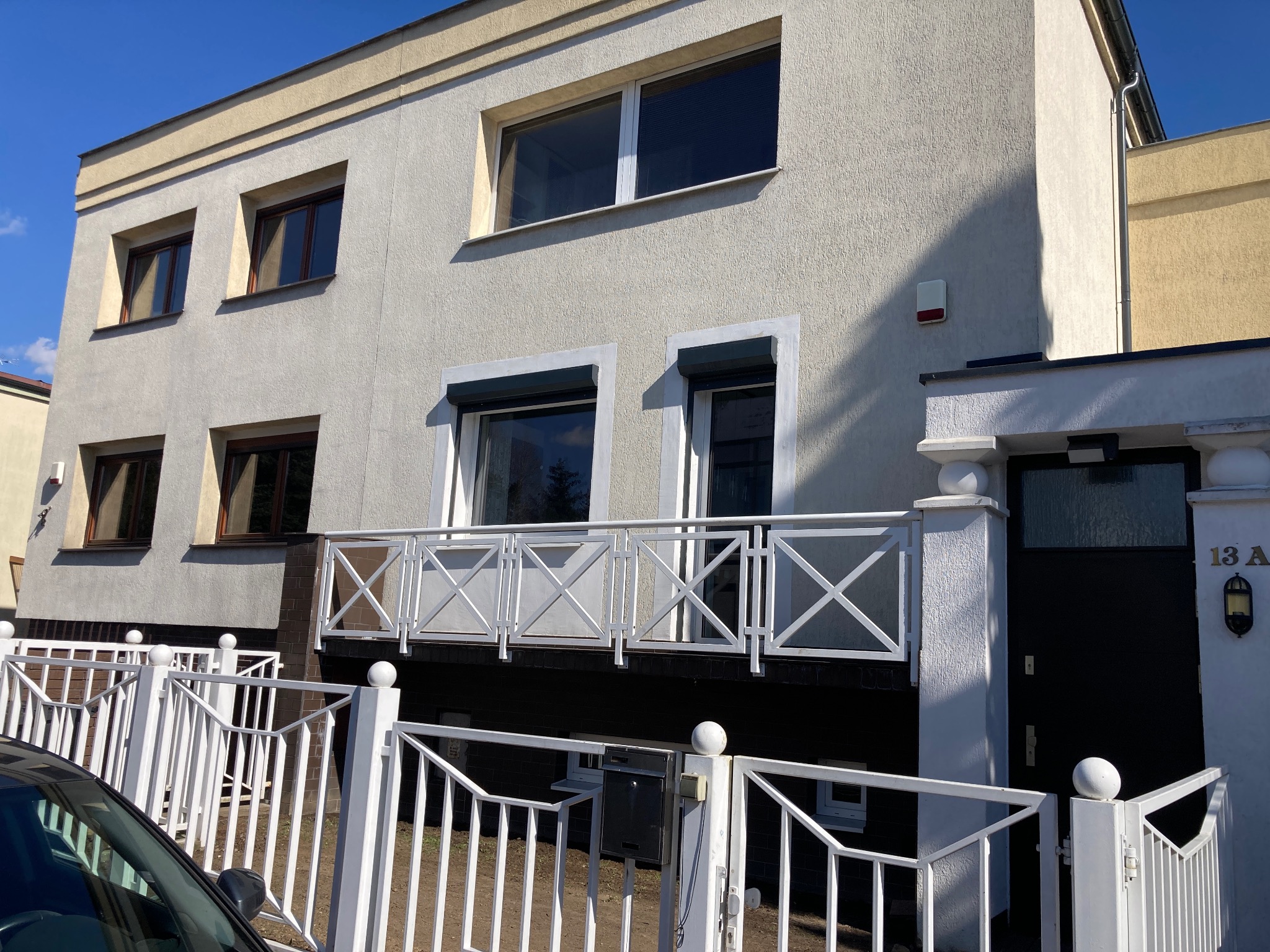 Dwupiętrowy budynek z jasną elewacją, balkonem z białą balustradą i białym ogrodzeniem, widziany z poziomu ulicy, z fragmentem samochodu w kadrze.