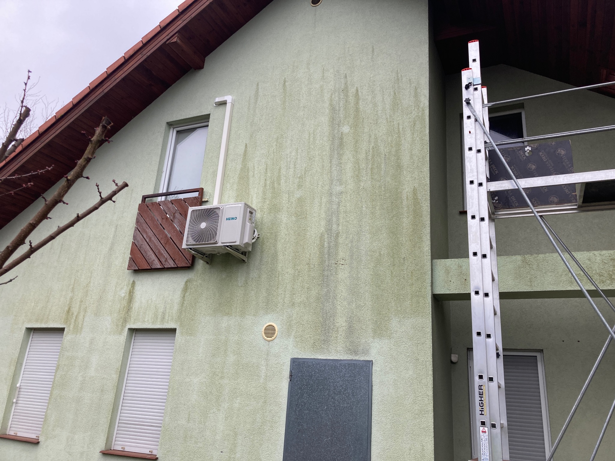 Brudna, zielonkawa elewacja domu z widocznymi zaciekami i zabrudzeniami, częściowo zasłonięta przez rozstawioną aluminiową drabinę z podestem roboczym, okna z opuszczonymi roletami, brązowy dach...