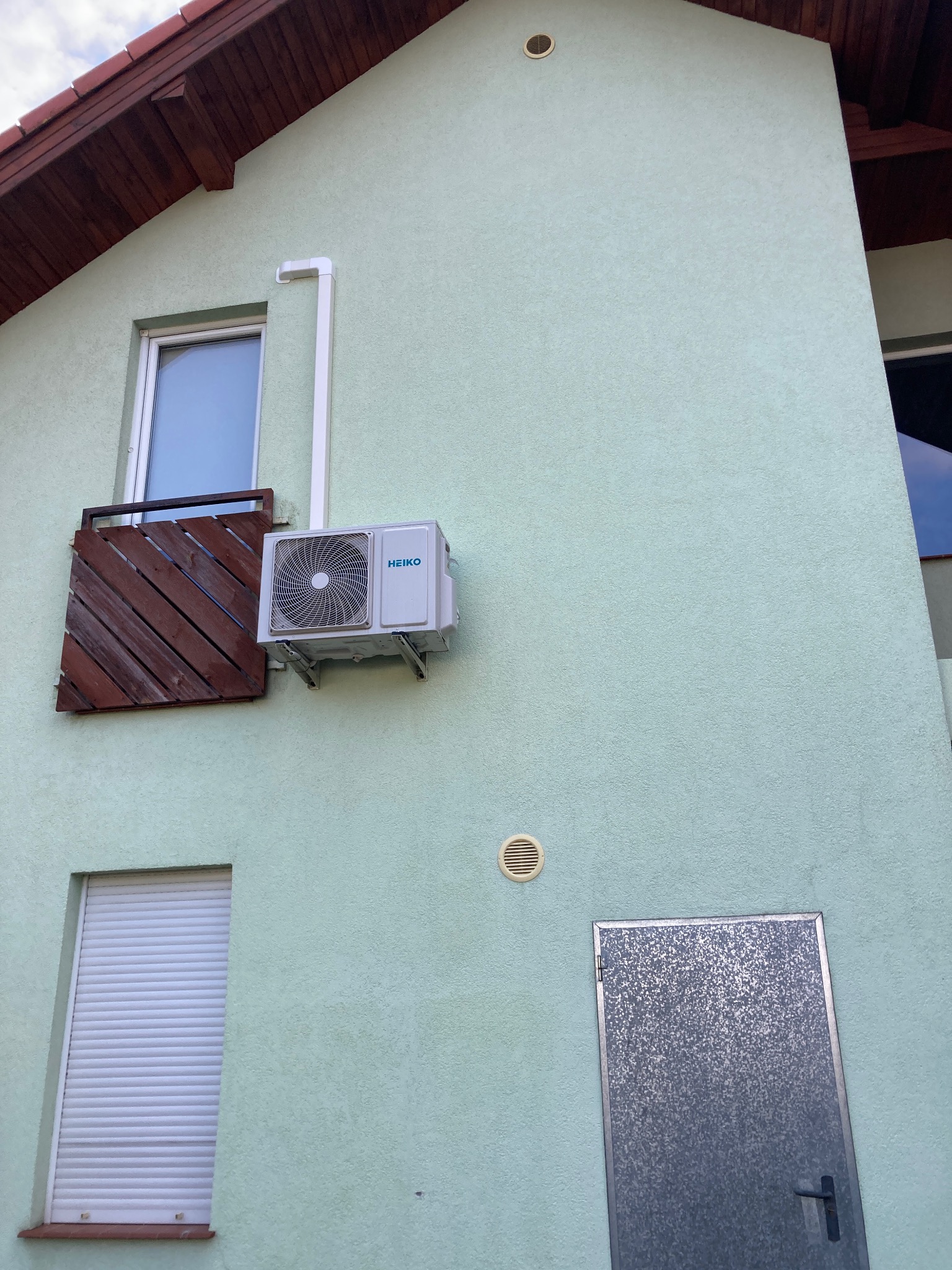 Jasnozielona elewacja budynku z zamontowanym klimatyzatorem marki Heiko, widoczne okna z roletami i drewnianą barierką, oraz metalowe drzwi.