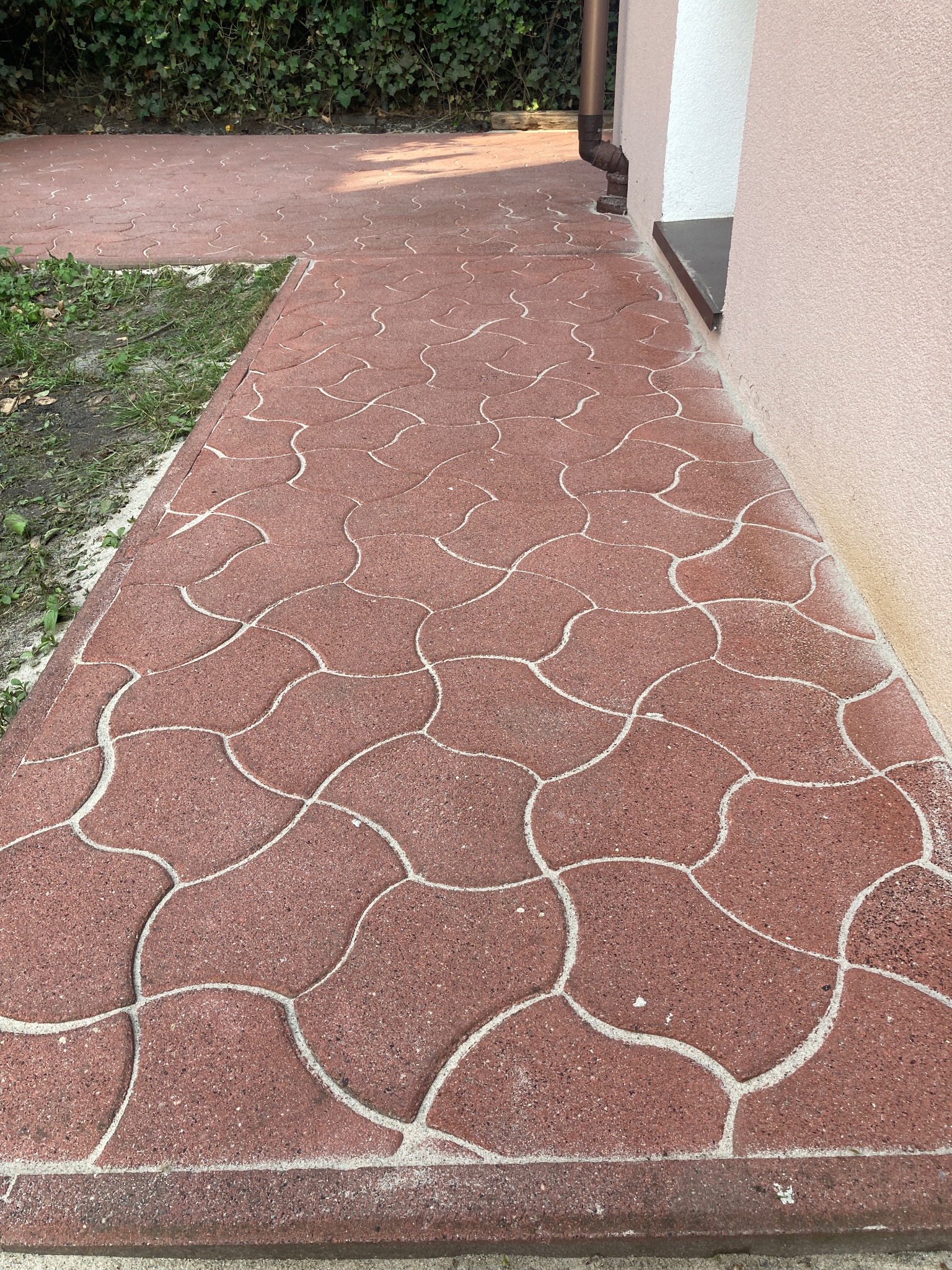 Czerwona kostka brukowa ułożona w falisty wzór prowadząca wzdłuż ściany budynku z widocznym systemem rynnowym.