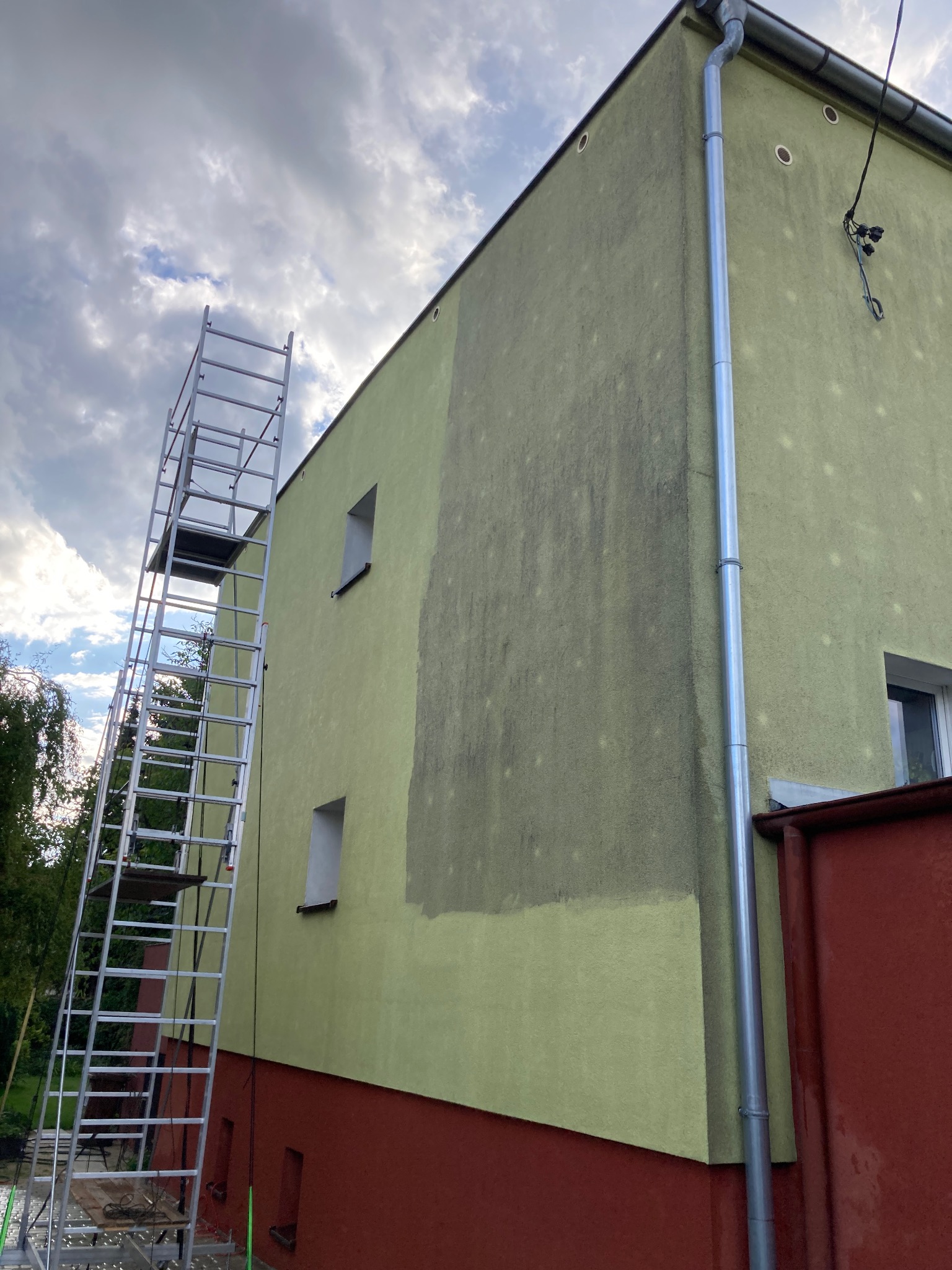 Fragment elewacji budynku z widocznym efektem mycia pod ciśnieniem, kontrast między umytą a brudną powierzchnią, aluminiowa drabina przystawiona do ściany, rynna pionowa.