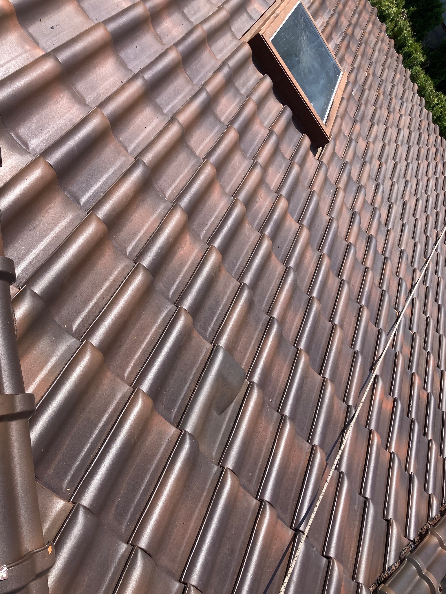 Błyszcząca, brązowa dachówka ceramiczna ułożona na dachu z oknem połaciowym. Widoczna lina zabezpieczająca. Ujęcie z góry, w słoneczny dzień.