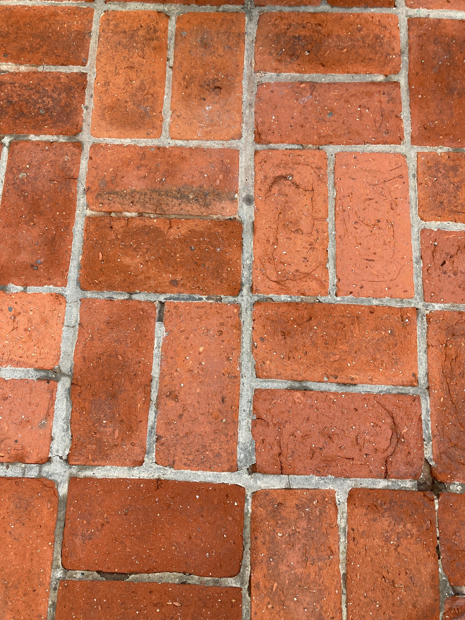 Ceglana kostka brukowa z szarymi fugami, widziana z góry. Czerwono-pomarańczowe cegły tworzą regularny wzór. Widoczne zabrudzenia i ślady użytkowania.