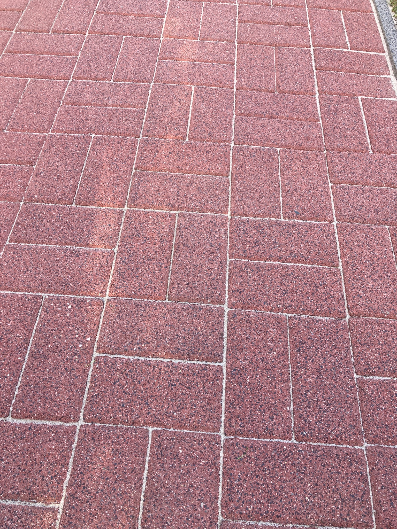 Czerwona kostka brukowa ułożona w regularny wzór. Widoczne fugi wypełnione piaskiem. Fragment trawnika w prawym górnym rogu.