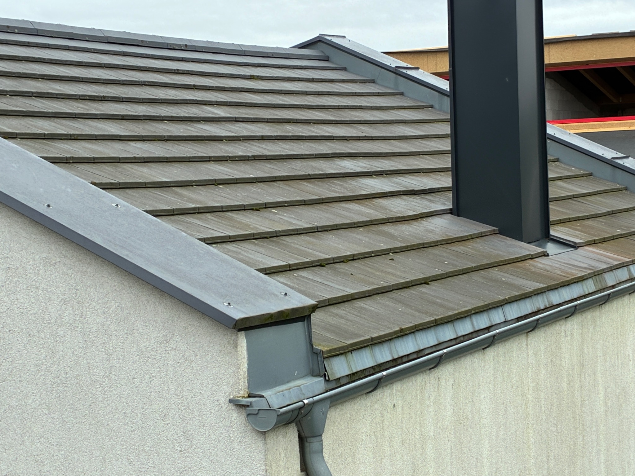 Ciemna dachówka ceramiczna na dachu domu jednorodzinnego z kominem i systemem rynnowym w kolorze szarym. Widoczny fragment elewacji z szorstkim tynkiem.