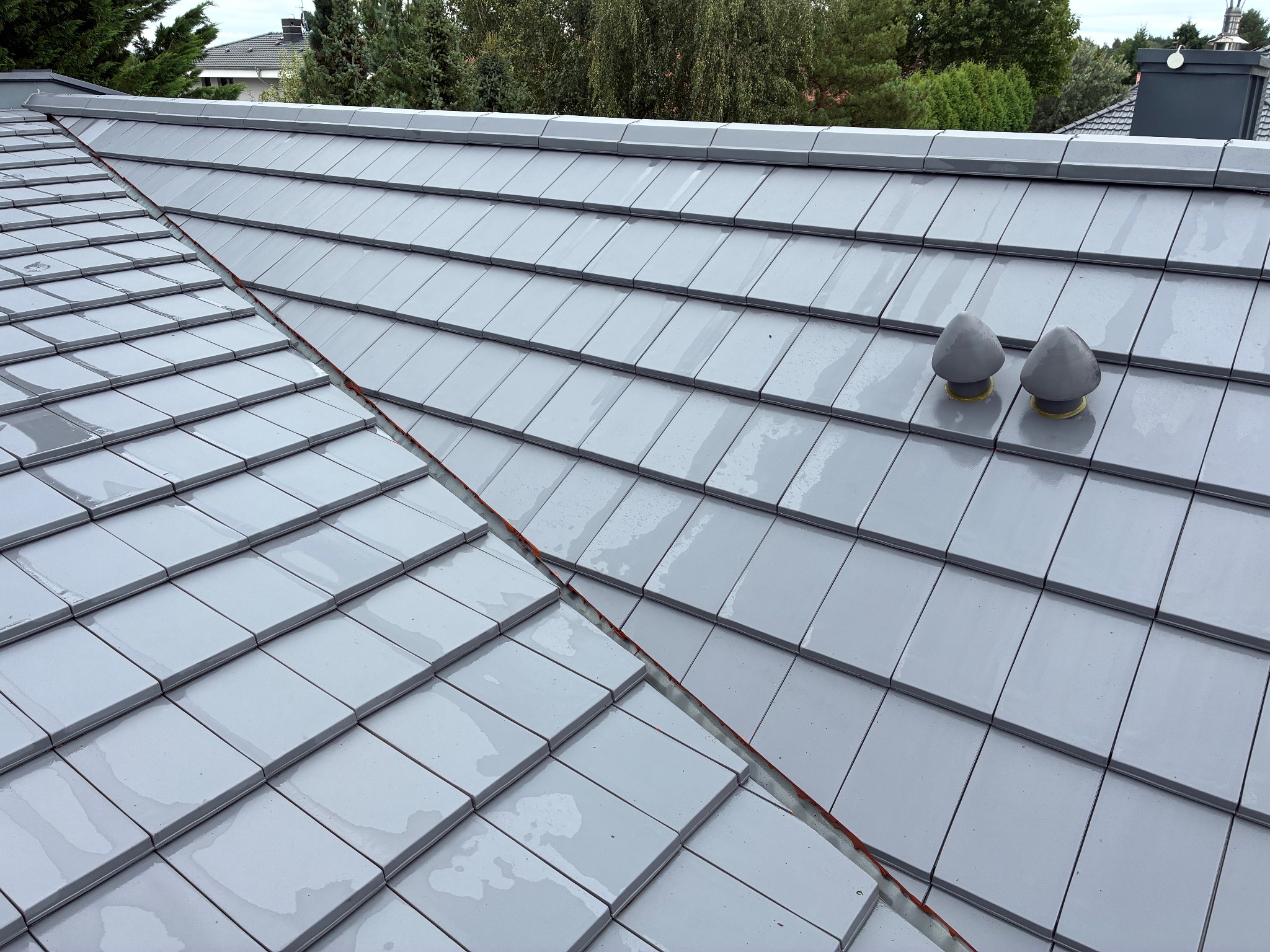 Błyszczący, szary dach z dachówki ceramicznej, widok z góry po deszczu, z dwoma odpowietrznikami kominowymi w kształcie grzybków.