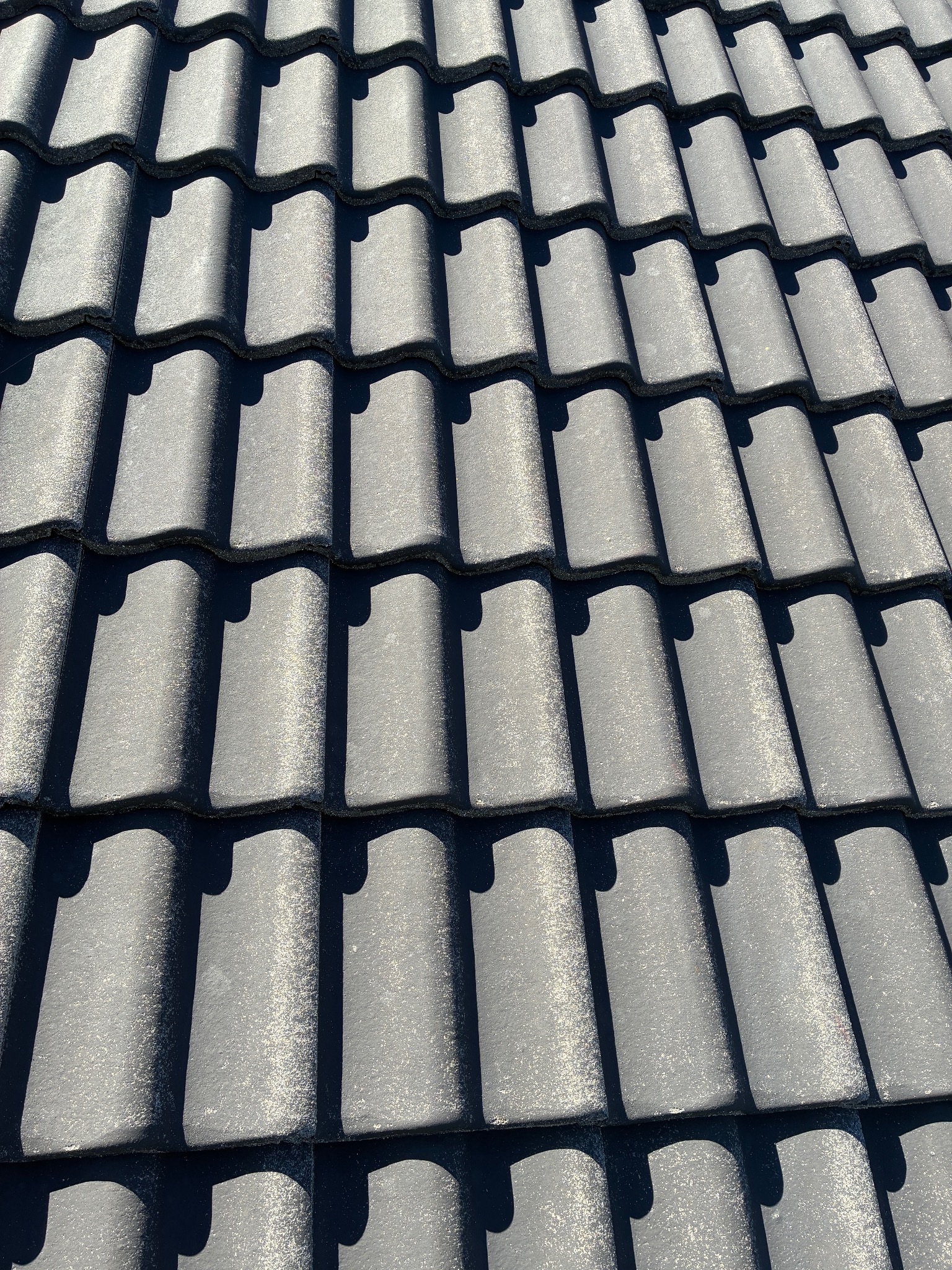 Czyste, ciemnoszare dachówki ceramiczne w rzędach, oświetlone słońcem, tworzące wzór cieni. Widok z góry, ukazujący regularność i estetykę pokrycia dachowego.