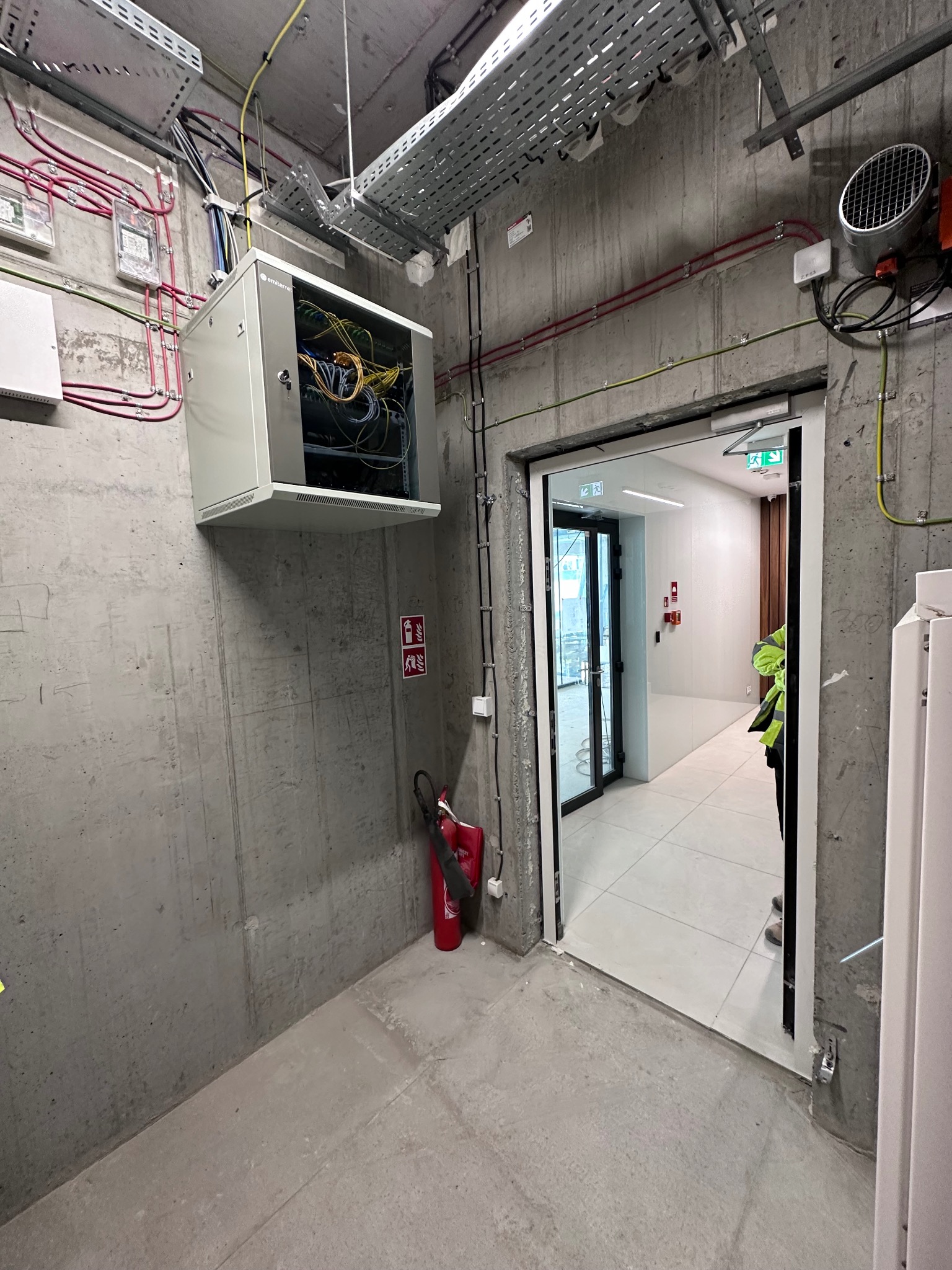 Szara szafa rack z okablowaniem zamontowana na betonowej ścianie w korytarzu biurowca. Widoczne kable, przewody i elementy instalacji elektrycznej.