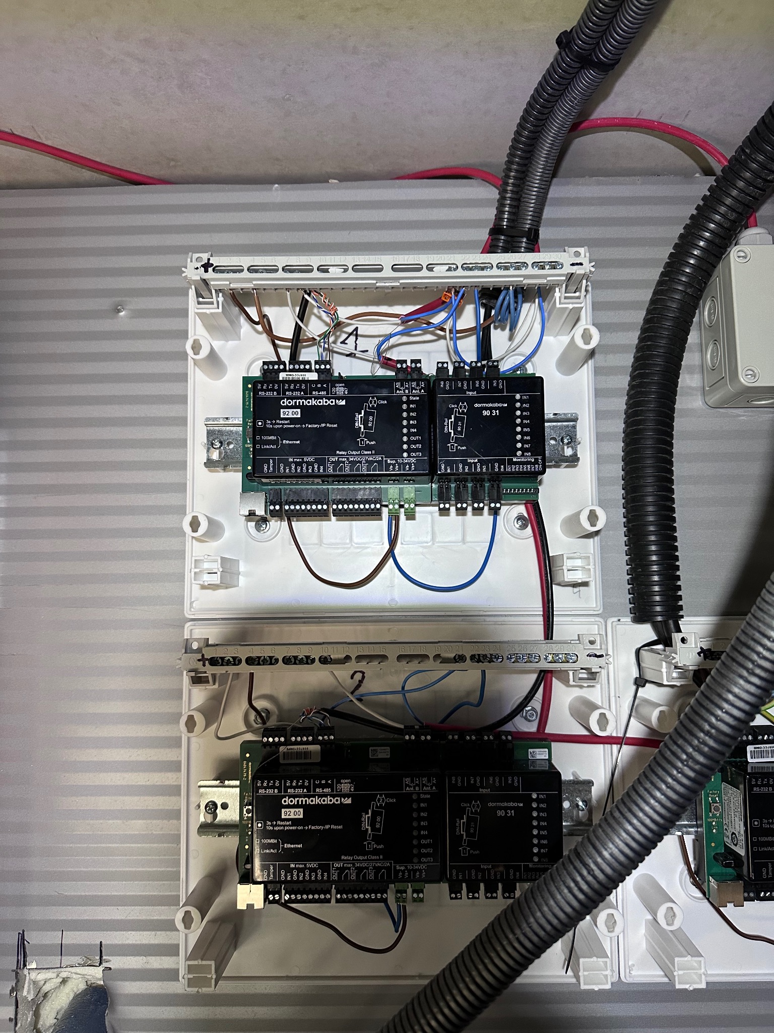 Dwa białe panele sterowania Dormakaba 92.00 i 90 31 z podłączonymi kablami, zamontowane na ścianie. Widoczne przewody w peszlach.