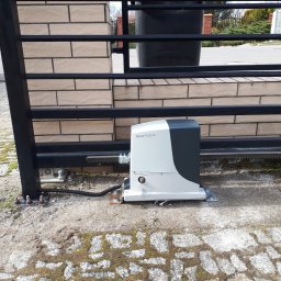 Przedsiębiorstwo Handlowo Usługowe Mariusz Mażul - Automatyka bramy przesuwnej Nice Robus zainstalowana przy czarnej, metalowej bramie z beżowym murowanym słupkiem, widoczne elementy montażowe na betonowej kostce z mchem.