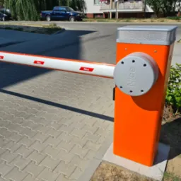 Pomarańczowo-szara szlaban automatyczny Nice na kostce brukowej, z widocznym logo, w tle budynki i samochody.