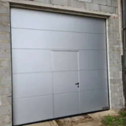 Zamknięta segmentowa brama garażowa w kolorze srebrnym z drzwiami przejściowymi i czarną klamką, zamontowana w surowym, betonowym otworze budynku.