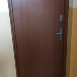 Brązowe drzwi wejściowe z wizjerem i srebrną klamką, zamontowane w ścianie pomalowanej na kremowo i żółto, z szarą podłogą wyłożoną płytkami.