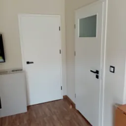 Dwie białe drzwi wewnętrzne z czarnymi klamkami w minimalistycznym stylu, jedna z małym przeszkleniem, na tle drewnopodobnej podłogi.