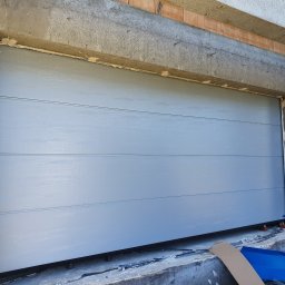 Przedsiębiorstwo Handlowo Usługowe Mariusz Mażul - Zamknięta, segmentowa brama garażowa w kolorze szarym, w surowym otoczeniu betonowych ścian i cegieł. Widoczne niedokończone wykończenia i elementy montażowe.