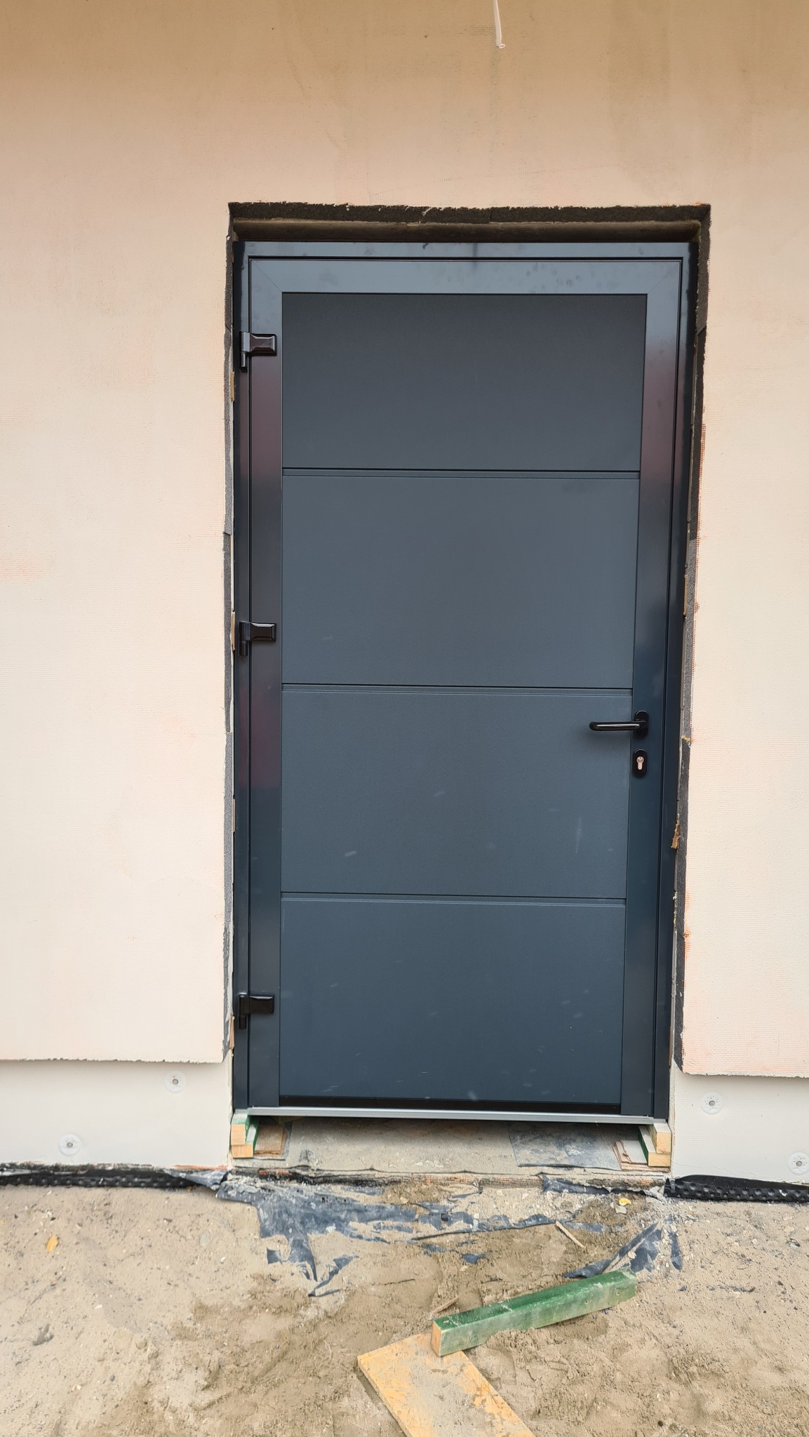 Szara brama garażowa segmentowa z poziomymi przetłoczeniami, zamontowana w surowym otworze budynku w trakcie prac wykończeniowych elewacji.