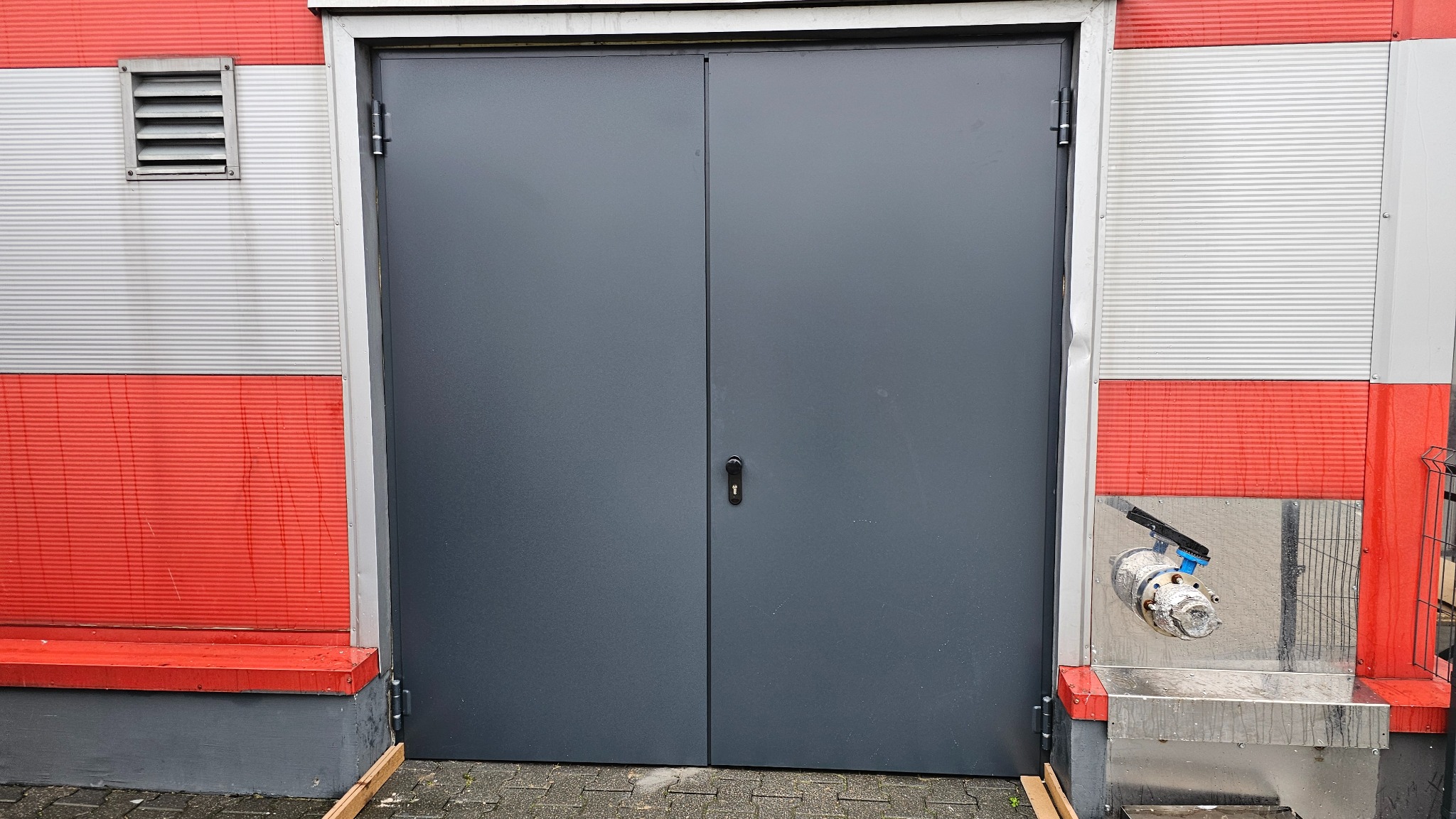 Szara, dwuskrzydłowa brama na tle czerwono-białej ściany z blachy falistej, z widoczną instalacją wodną po prawej stronie.