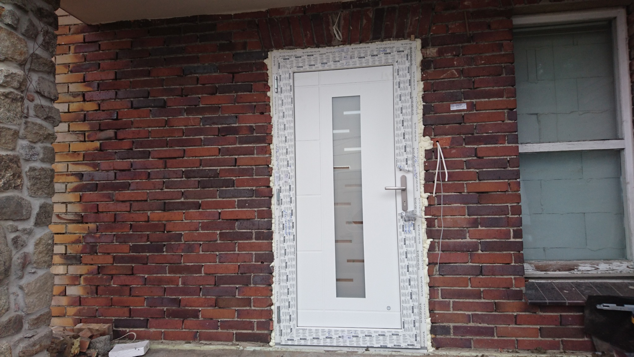 Białe drzwi z szybą w trakcie montażu, zabezpieczone pianką montażową, na tle ściany z czerwonej cegły i okna z zasłoniętą szybą.