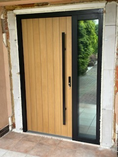 Nowoczesne drzwi wejściowe z panelem drewnianym i bocznym przeszkleniem, czarna rama, widoczny fragment elewacji w trakcie wykończenia.