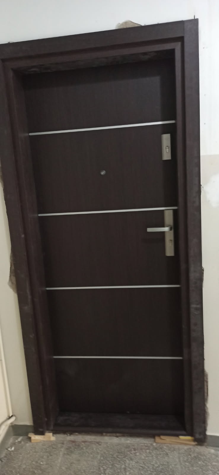 Ciemnobrązowe drzwi wejściowe z czterema poziomymi, srebrnymi paskami, wizjerem, zamkiem i klamką, osadzone w ciemnej futrynie, widoczne drewniane kliny pod futryną.