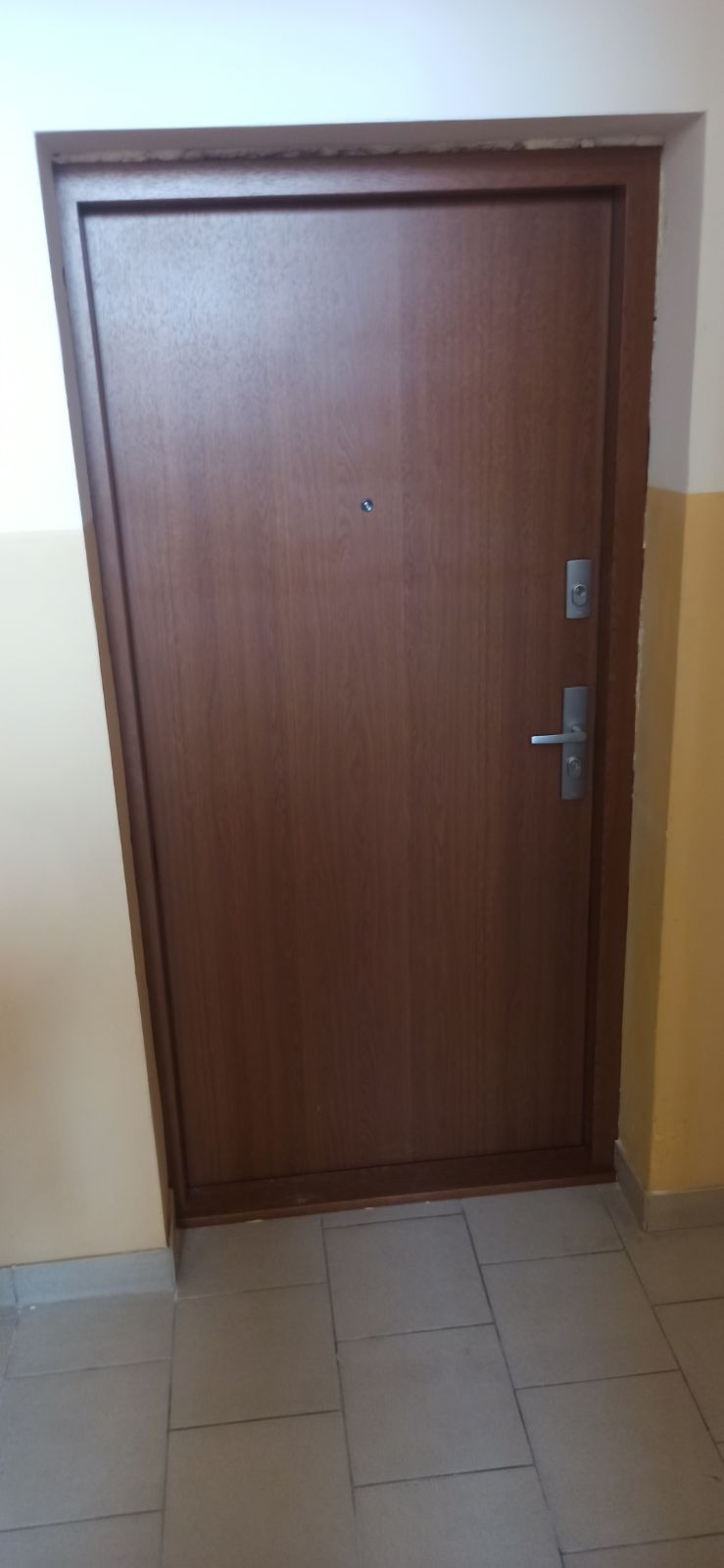 Brązowe drzwi wejściowe z wizjerem i srebrną klamką, zamontowane w ścianie pomalowanej na kremowo i żółto, z szarą podłogą wyłożoną płytkami.