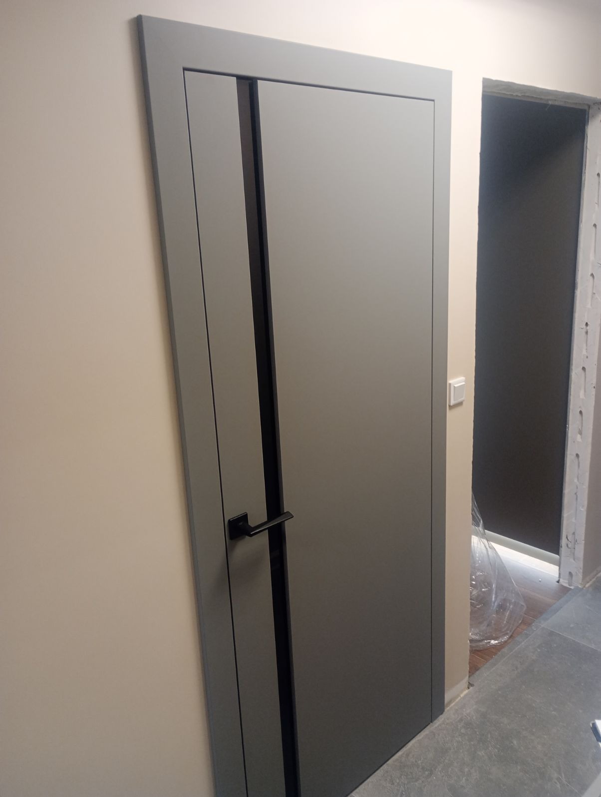 Szare drzwi wewnętrzne z czarnym pionowym akcentem i klamką, widoczne w trakcie montażu, z folią ochronną na podłodze w przejściu.