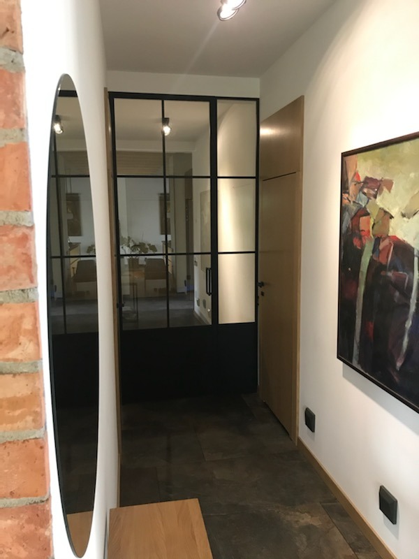 Nowoczesne wnętrze przedpokoju z czarną, przeszkloną konstrukcją oddzielającą strefy, owalne lustro na ceglanej ścianie i abstrakcyjny obraz na białej ścianie.