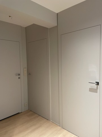 Trzy wewnętrzne drzwi drewniane w odcieniach szarości, z minimalistycznymi srebrnymi klamkami, wkomponowane w szarą ścianę i jasną drewnianą podłogę.