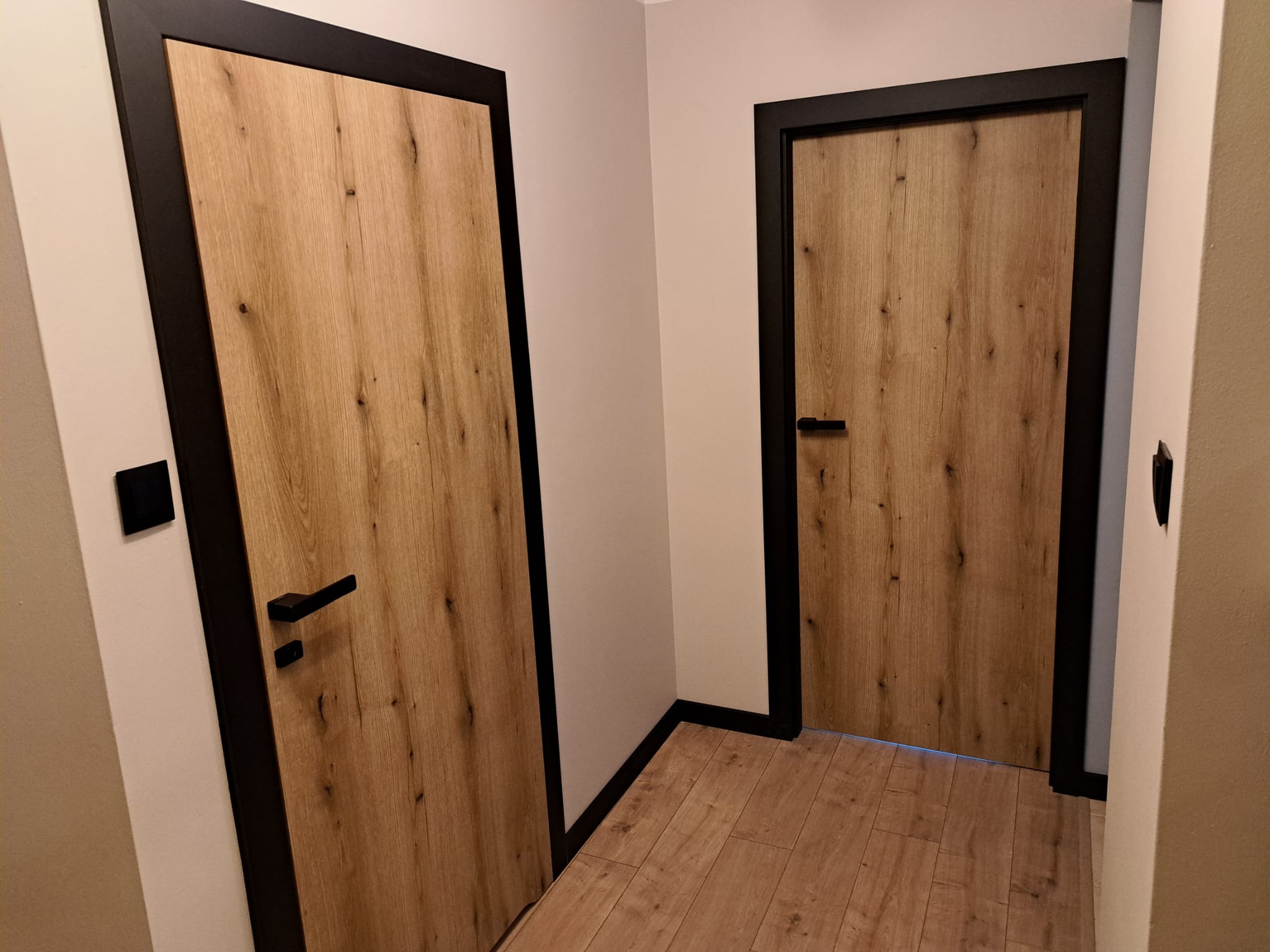Dwa skrzydła drzwiowe z okleiną imitującą jasne drewno, czarne ościeżnice i klamki, widok z niskiej perspektywy, podłoga z paneli w odcieniach drewna.
