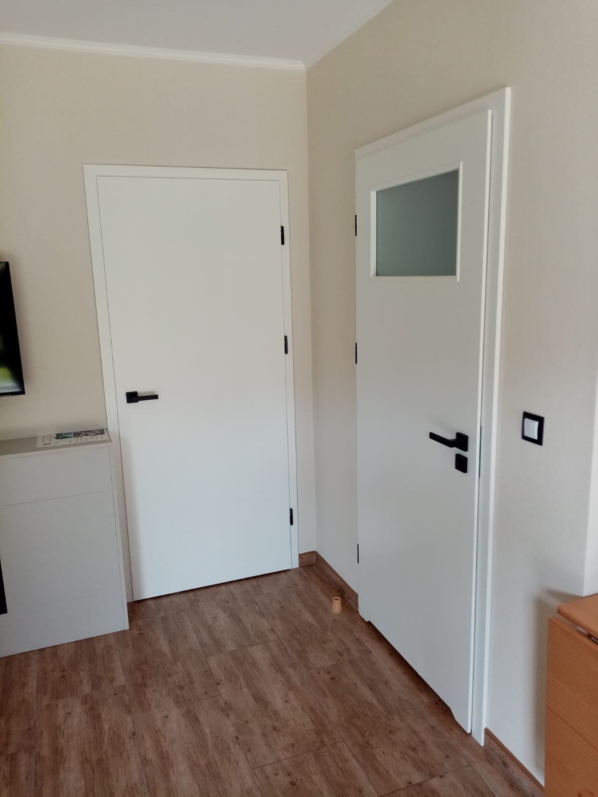 Dwie białe drzwi wewnętrzne z czarnymi klamkami w minimalistycznym stylu, jedna z małym przeszkleniem, na tle drewnopodobnej podłogi.