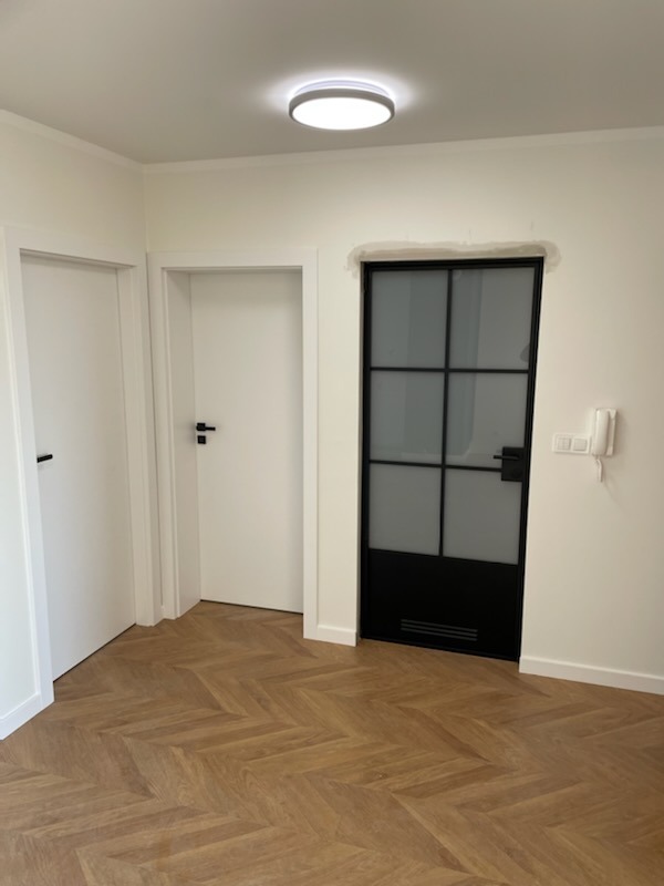 Wnętrze z trzema drzwiami: dwoma białymi i jednymi czarnymi, szklanymi w metalowej ramie, parkiet ułożony we wzór jodełki, białe ściany i sufit z okrągłą lampą.