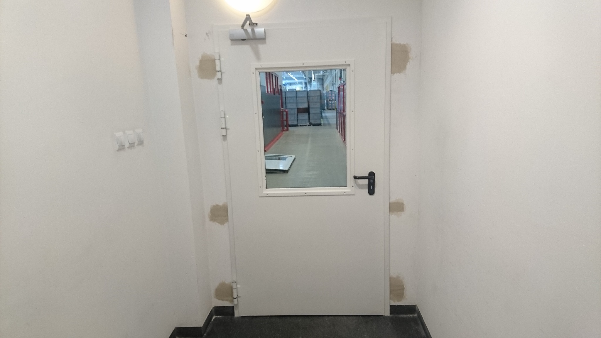 Białe, stalowe drzwi z małym okienkiem w magazynie, ściany z widocznymi śladami po szpachlowaniu.