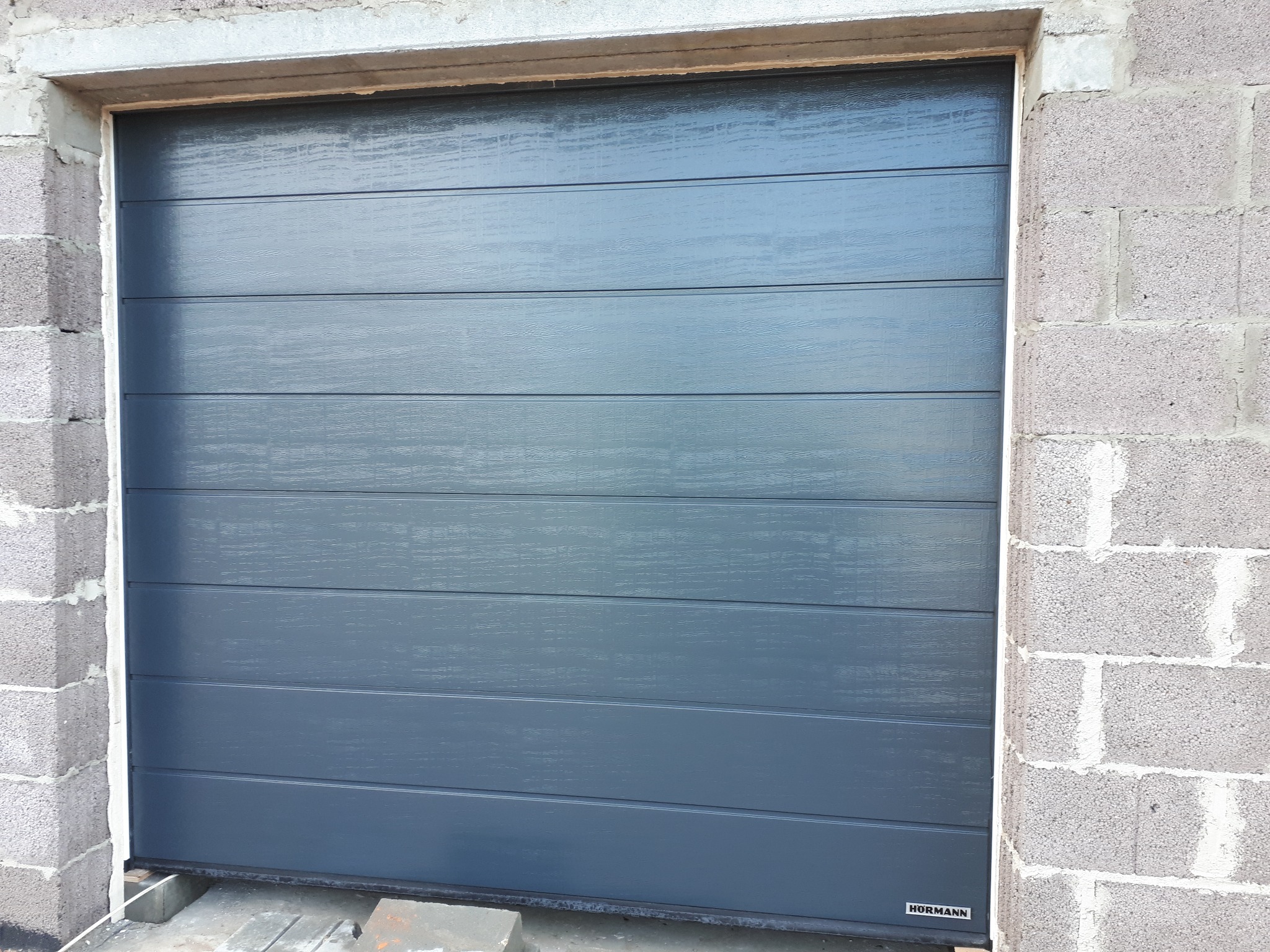 Zamknięta, segmentowa brama garażowa Hormann w odcieniach szarości, widoczna z bliska na tle surowych, betonowych bloczków ściany garażu.
