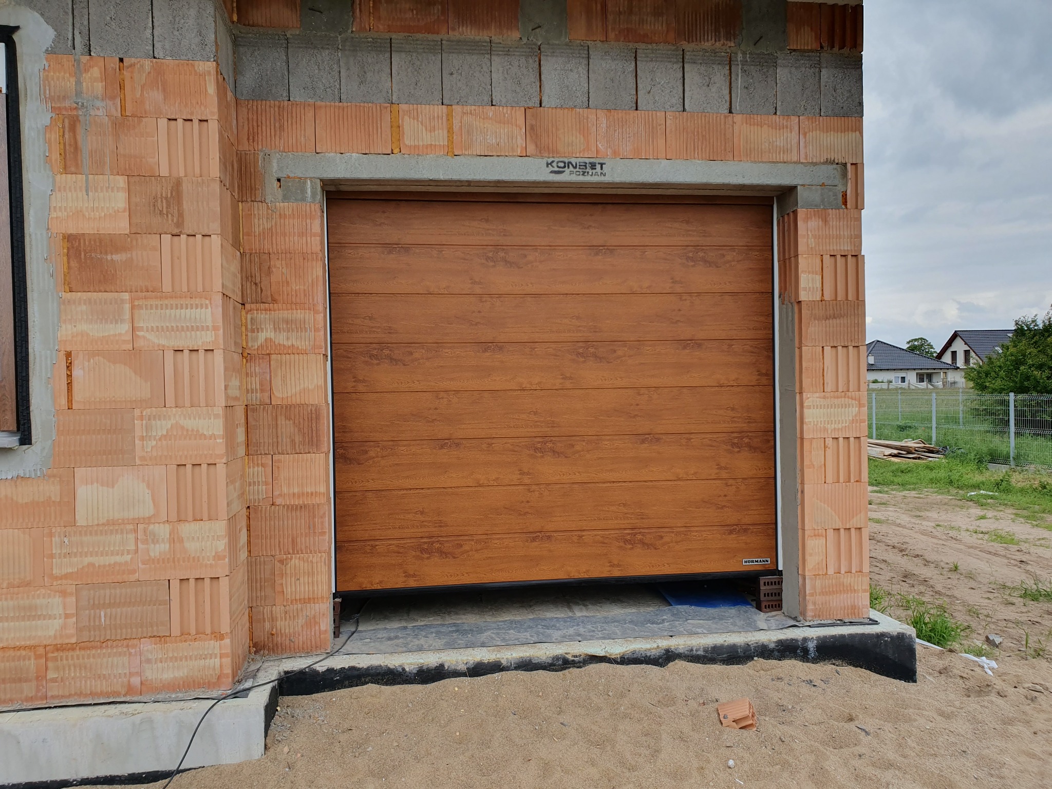 Zamknięta brama garażowa segmentowa w kolorze drewna, w surowym otworze budynku z cegły. Widoczna marka Konbet Poznań i Hormann. Tło: budynki mieszkalne.