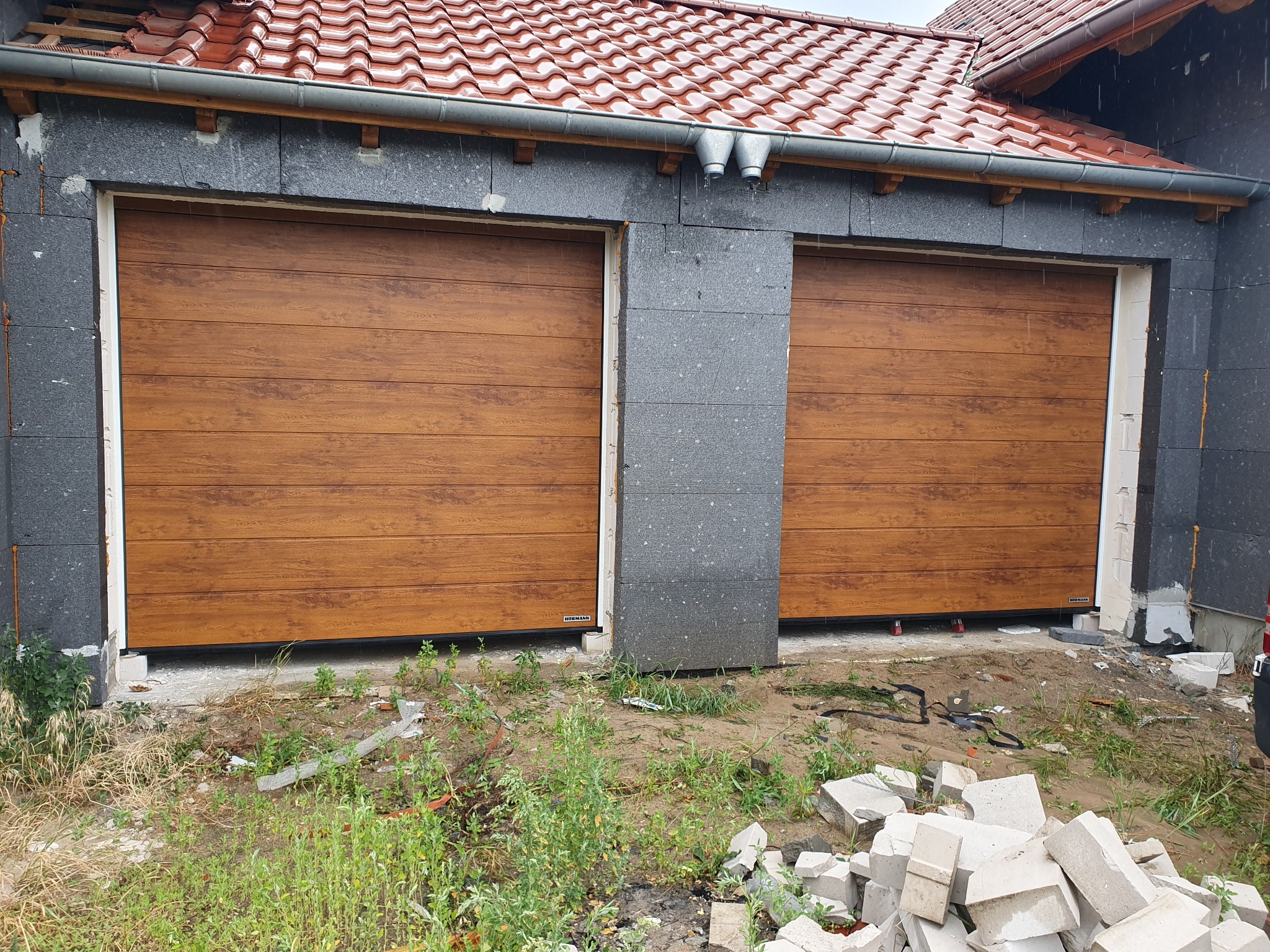 Dwie brązowe bramy garażowe segmentowe, z widoczną strukturą drewna, zamontowane w surowym otoczeniu, z elementami budowlanymi na ziemi.