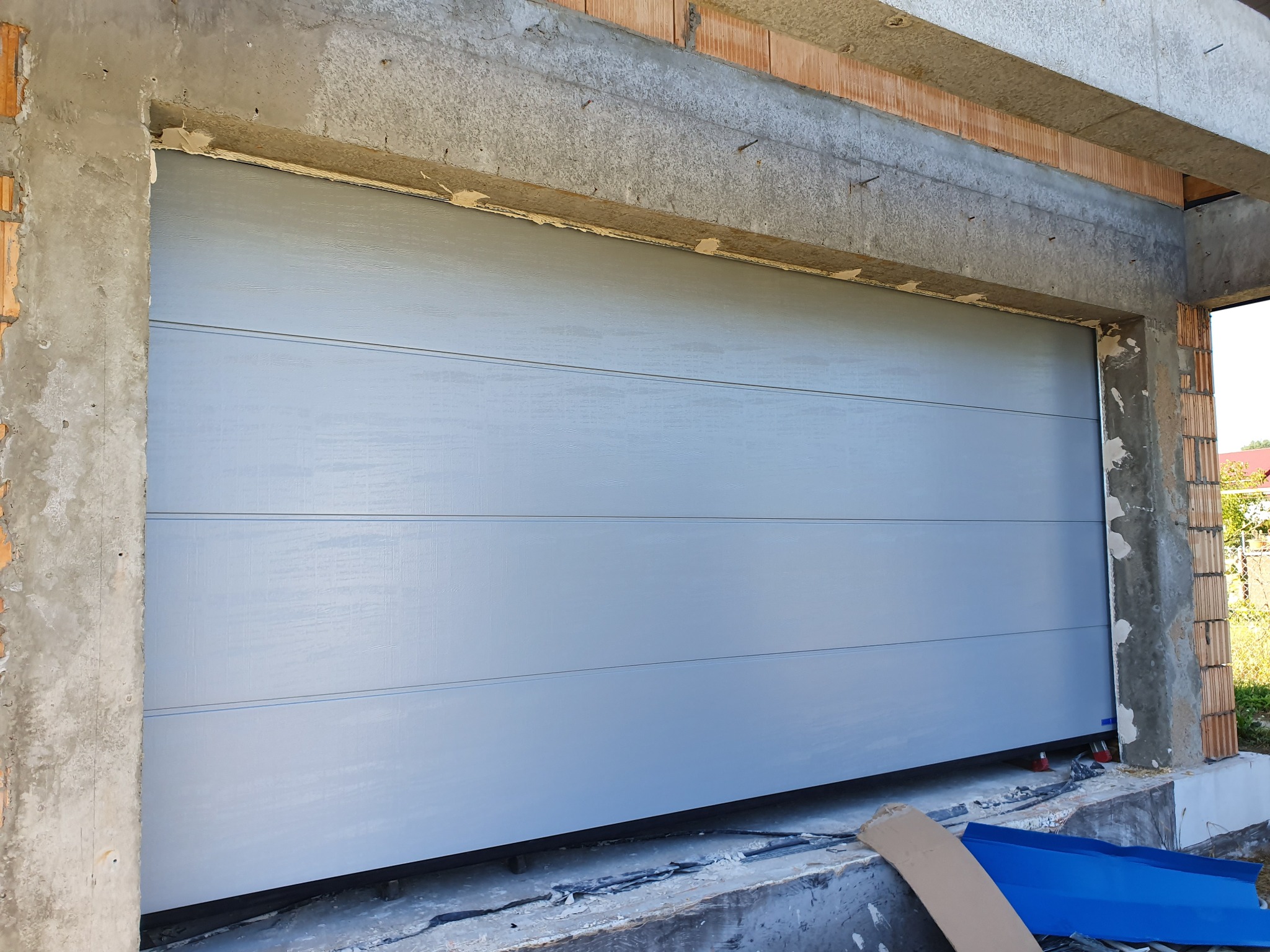 Zamknięta, segmentowa brama garażowa w kolorze szarym, w surowym otoczeniu betonowych ścian i cegieł. Widoczne niedokończone wykończenia i elementy montażowe.