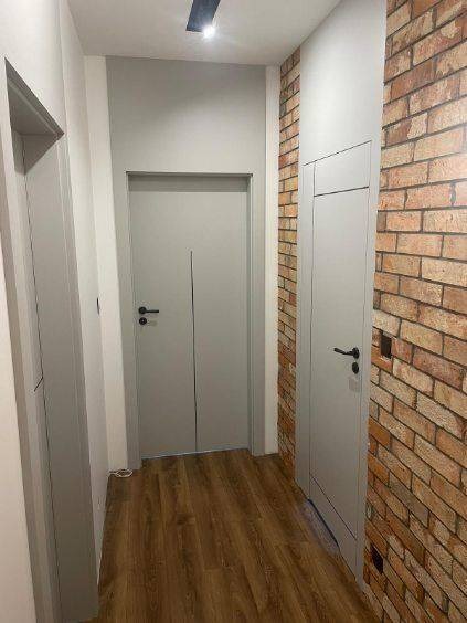 Szare, minimalistyczne drzwi wewnętrzne z czarnymi klamkami, wkomponowane w nowoczesne wnętrze z akcentem ceglanej ściany i drewnianej podłogi.