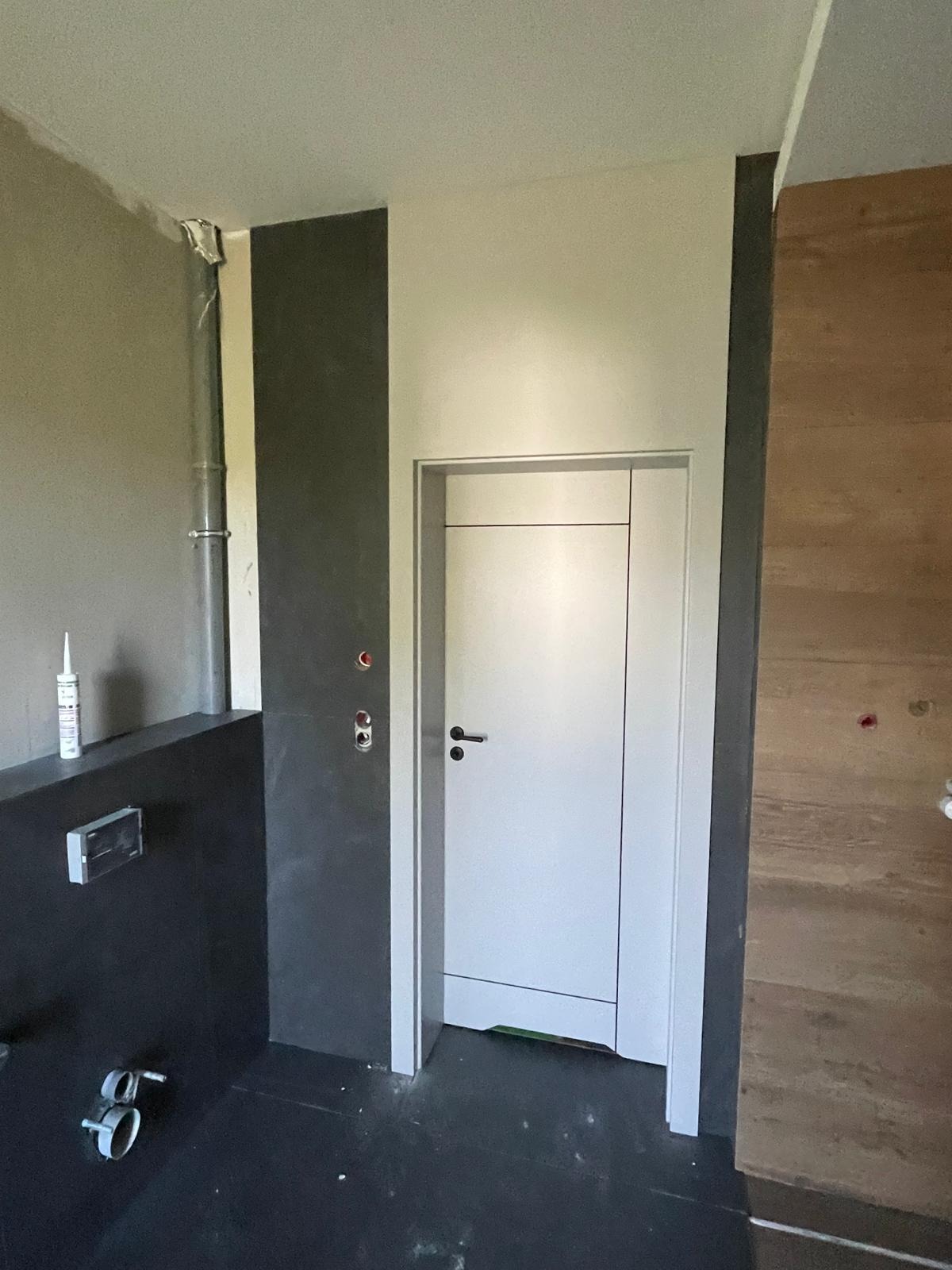 Białe drzwi wewnętrzne z czarną klamką w surowym wnętrzu. Widoczna instalacja sanitarna i fragment ściany wykończonej drewnem. Minimalistyczny design.