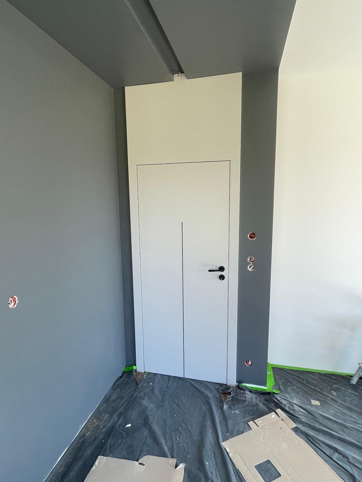 Białe, minimalistyczne drzwi wewnętrzne z czarną klamką, wkomponowane w szarą ścianę. Widoczne zabezpieczenia podłogi folią malarską.