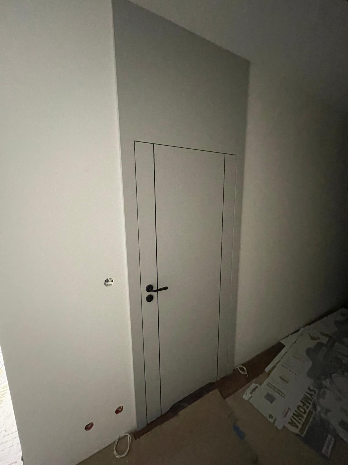 Białe drzwi wewnętrzne z czarną klamką, minimalistyczny design, widoczne ślady prac wykończeniowych na ścianie i podłodze. Ujęcie z bliska, podkreślające detale.