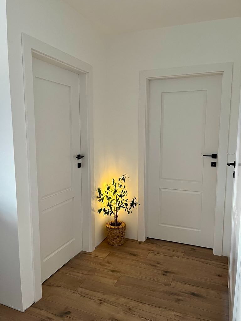 Dwa białe, minimalistyczne skrzydła drzwiowe z czarnymi klamkami, ustawione w korytarzu. Pomiędzy nimi roślina w plecionym koszu, oświetlona ciepłym światłem.