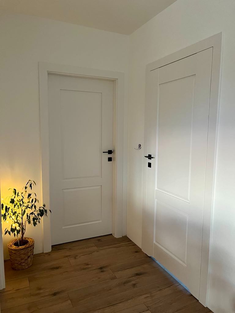 Dwa białe, panelowe skrzydła drzwiowe z czarnymi klamkami w jasnym wnętrzu. Podłoga z paneli imitujących drewno, roślina w wiklinowym koszu z lewej strony.