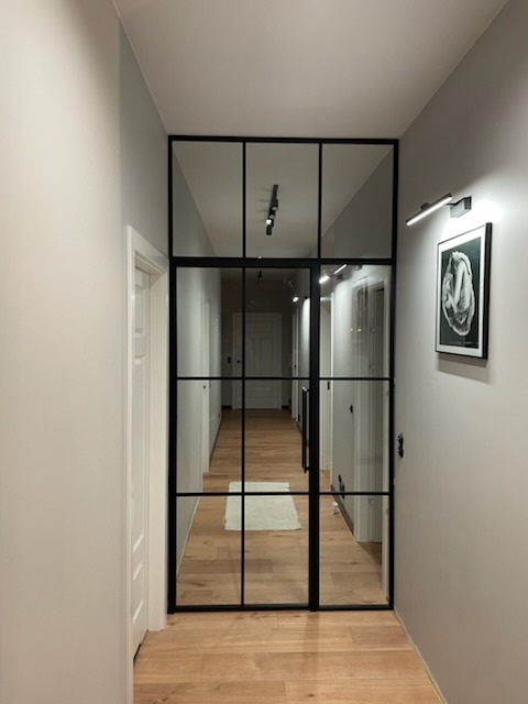 Szklane drzwi wewnętrzne z czarnymi szprosami, oddzielające korytarz z drewnianą podłogą. Widok na dalszą część korytarza, oświetlenie punktowe.