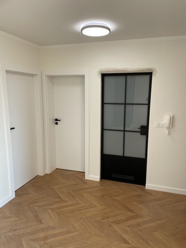 Nowoczesne wnętrze z trzema parami drzwi: dwie białe, klasyczne i jedne czarne, loftowe z szybą, na podłodze parkiet w jodełkę. Minimalistyczny design.