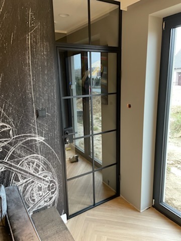 Nowoczesne drzwi wewnętrzne z czarną ramą i szprosami, widok na zewnątrz, parkiet w jodełkę, ściana z motywem graficznym roweru. Minimalistyczny design.